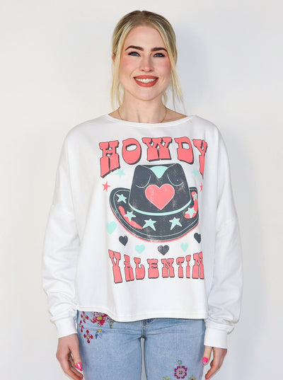 Model is wearing a white fleece sweatshirt that reads "Howdy Valentine" in a bold pink text with a large cowboy hat with pink, blue, and black star and heart details. Sweatshirt is paired with blue jeans.