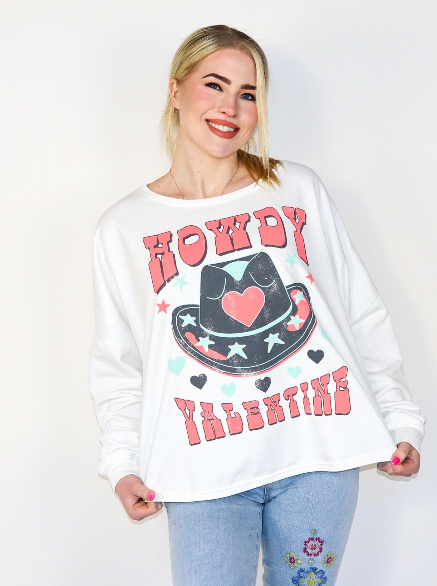 Model is wearing a white fleece sweatshirt that reads "Howdy Valentine" in a bold pink text with a large cowboy hat with pink, blue, and black star and heart details. Sweatshirt is paired with blue jeans. 