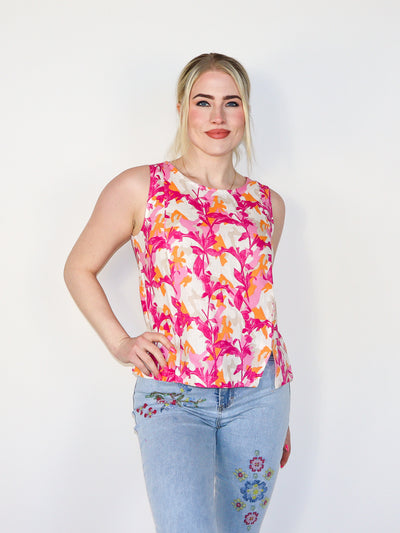 Model is wearing light pink, white, and hot pink leaf printed sleeveless top. Top has slits on the bottom on both sides. Worn with blue jeans. 