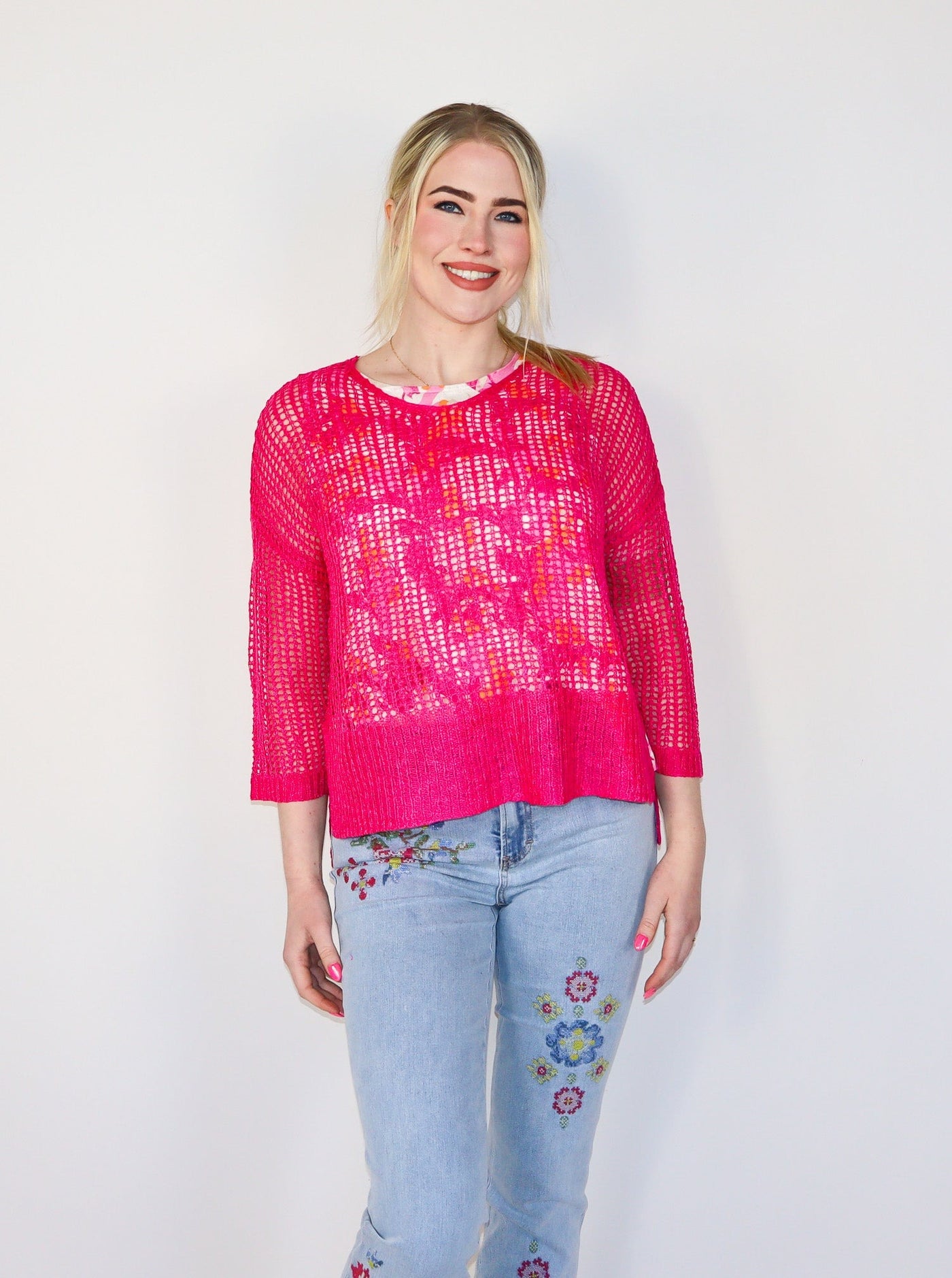 Model is wearing a hot pink fishnet crochet sweater. Sweater is paired with a floral tank top underneath and blue jeans. 