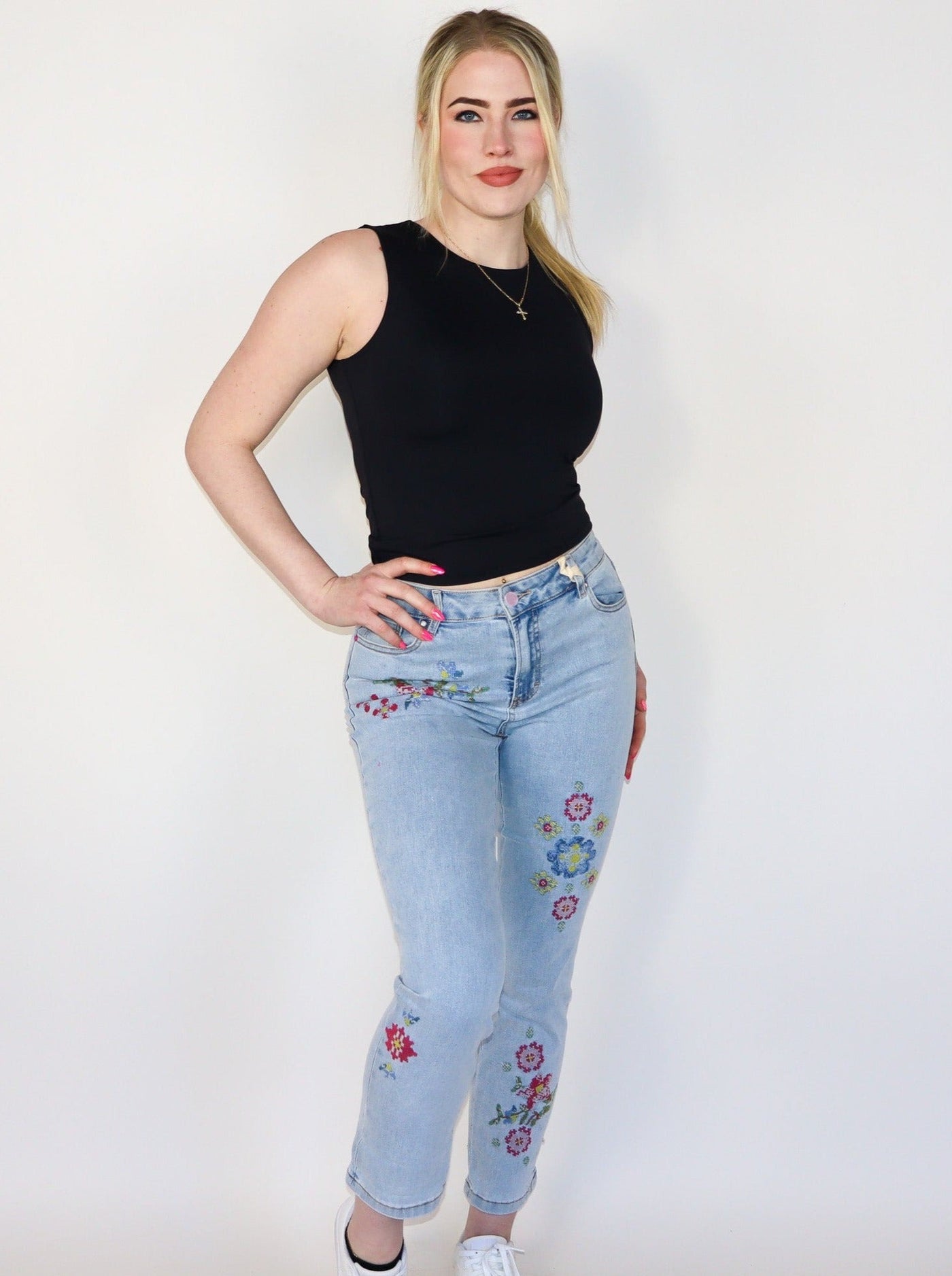 Model is wearing a light wash blue jean with floral stitching on the legs. Jeans are paired with a black tank top. 