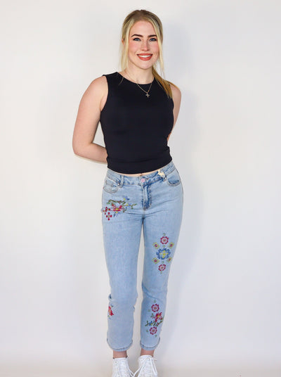 Model is wearing a light wash blue jean with floral stitching on the legs. Jeans are paired with a black tank top. 