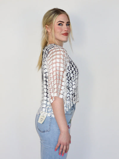 Model is wearing a white see through floral embroidered half sleeve top. Top is paired with a black tank top and blue jeans. 