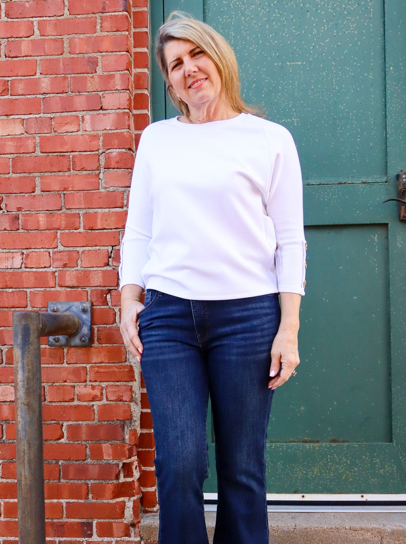 Model is wearing a white 3/4th sleeve top with brown buttons on the sleeve. Top is paired with blue jeans. 