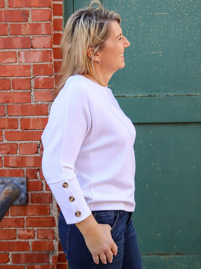 Model is wearing a white 3/4th sleeve top with brown buttons on the sleeve. Top is paired with blue jeans.