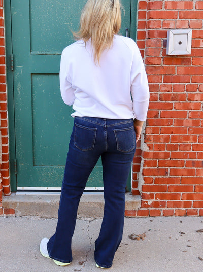 Model is wearing dark wash flare high rise blue jeans paired with a white top and white sneakers.