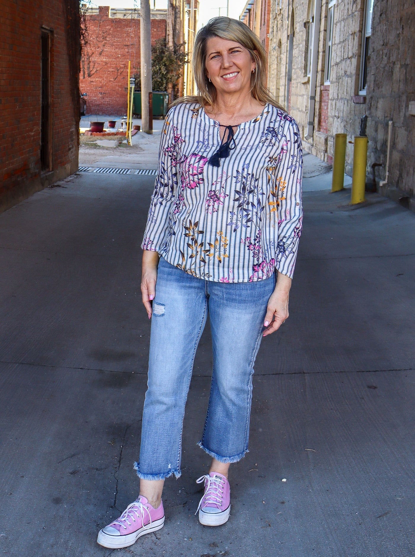 Model is wearing a vertical striped long sleeve blouse with multi color floral detail. The blouse has a string on the neckline for a bowtie detail. Blouse is paired with blue jeans and pink converse.