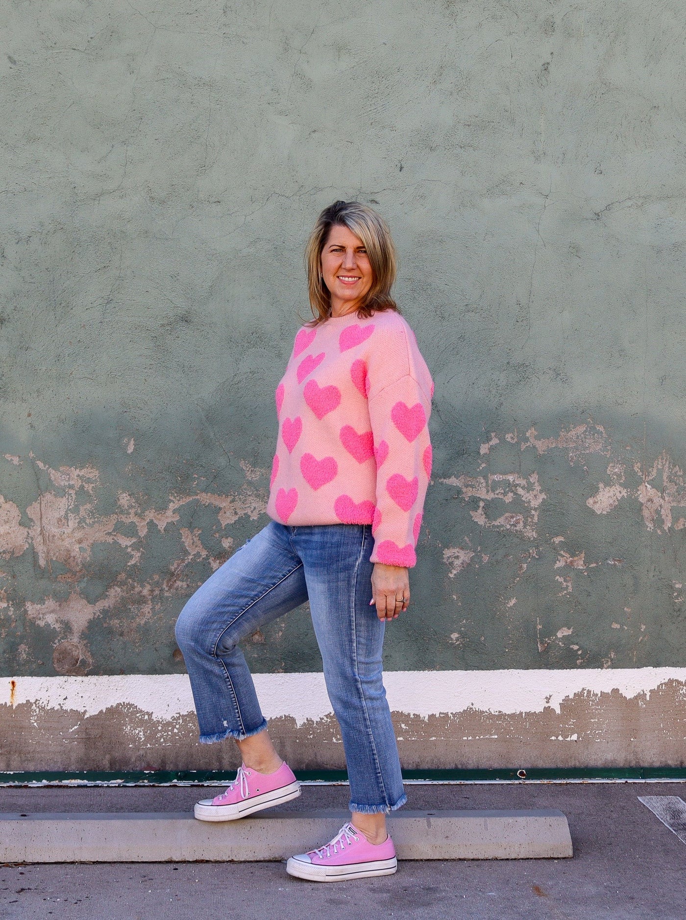 Model is wearing a peach and hot pink colored fuzzy sweater with heart details. Sweater is paired with blue jeans and pink converse. 
