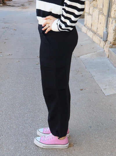 Model is wearing 3/4th length black cargo pants. Pants have 2 sets of pockets, one pair at waist and the other pair on the side, mid thigh. Cargo Pants are paired with a black and white sweater and pink converse. 