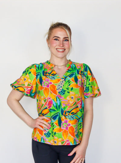Model is wearing a multi colored neon short sleeve blouse with a v neck and puffy sleeves. Top is paired with black pants. 