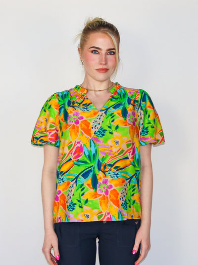 Model is wearing a multi colored neon short sleeve blouse with a v neck and puffy sleeves. Top is paired with black pants. 