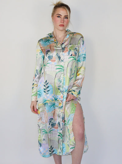 Model is wearing a long sleeve spring themed dress with multi color print. The dress buttons up in the front and has slits at the thighs on both sides. Dress is paired with white sneakers. 