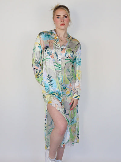 Model is wearing a long sleeve spring themed dress with multi color print. The dress buttons up in the front and has slits at the thighs on both sides. Dress is paired with white sneakers.