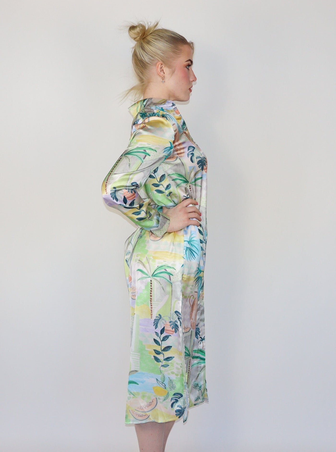 Model is wearing a long sleeve spring themed dress with multi color print. The dress buttons up in the front and has slits at the thighs on both sides. Dress is paired with white sneakers.