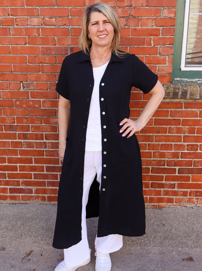 Model is wearing a black short sleeve duster that hits at the ankles and buttons up. Duster is paired with a white tank and white jeans. 