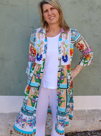 Model is wearing a multi colored printed duster. Duster is unbuttoned and paired with a white tank top and white jeans. Duster hits at the knee.