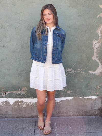 Model is wearing a dark wash blue jean jacket Jean jacket is worn with a white mini dress and sandals. 