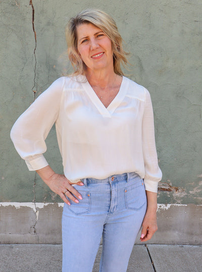 Model is wearing a long sleeve cream colored v-neck blouse. Paired with blue jeans.