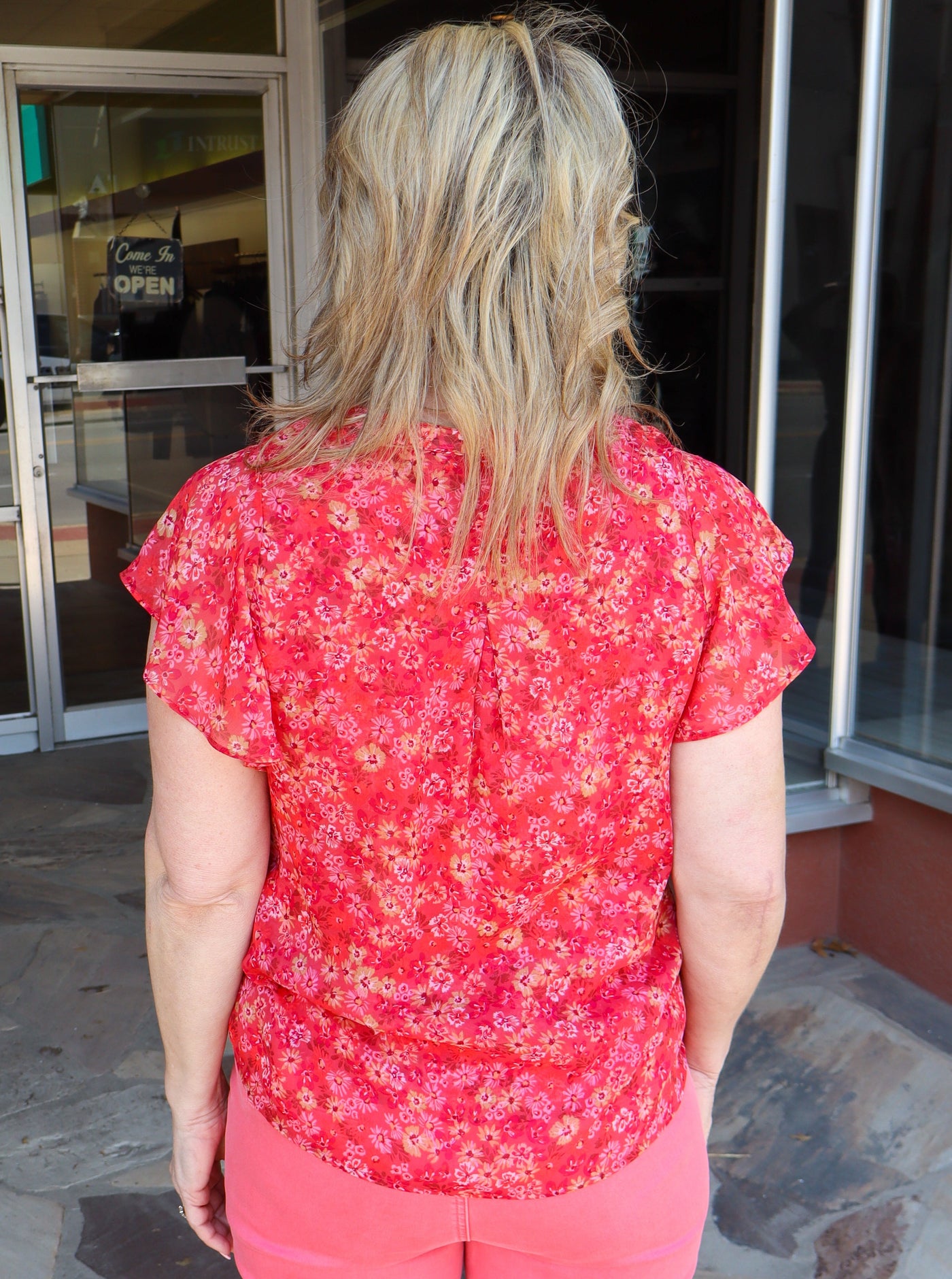 Model is wearing a rouched short sleeve coral colored floral blouse with flowy sleeve and a half button down the front.
