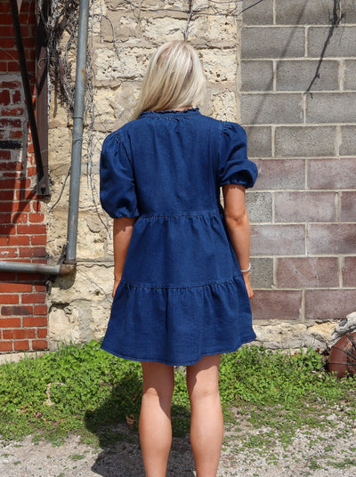 Model is wearing a dark wash denim babydoll style dress with tiering detail on the body, a slit neck, and puff sleeves.