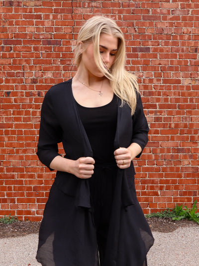 Model is wearing a long open flowy black half sleeve blouse. Paired with matching pants and sandals.