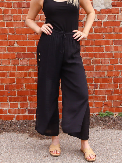 Model is wearing a black flowy pull on high waisted pant. Pants are doubled lined and have a unique slit with a double layer at each ankle. Pants have beige buttons that go up each side of hip. Pants are paired with a black tank and sandals.