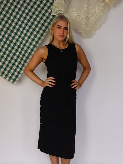Model is wearing a sleeveless black maxi dress with a scoop neck and silver snap buttons on each side of dress.