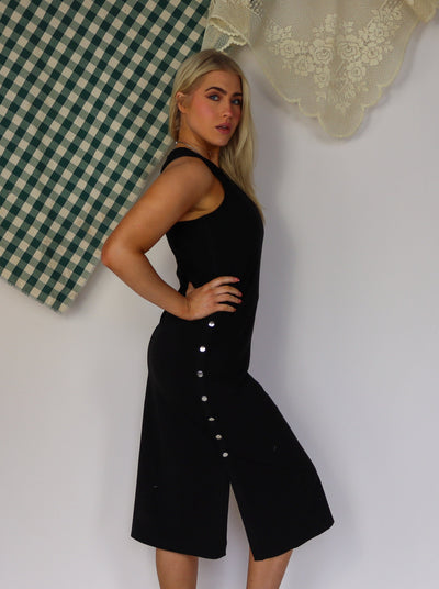 Model is wearing a sleeveless black maxi dress with a scoop neck and silver snap buttons on each side of dress.