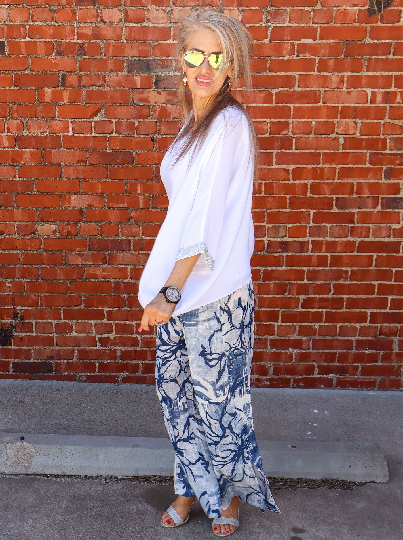 Model is wearing a white flowy blouse with sparkle at the neckline and cuffing of the sleeves. Top is paired with linen blue and white pants.