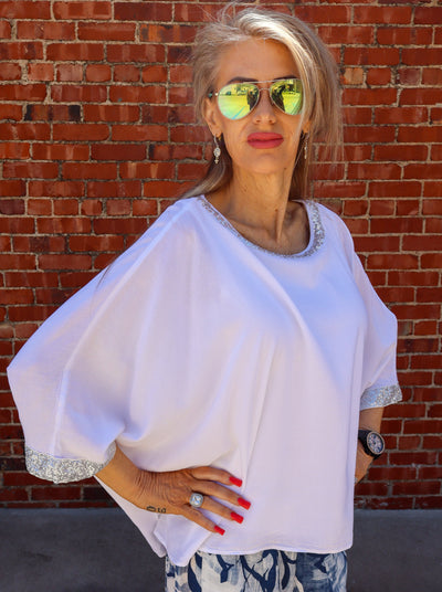 Model is wearing a white flowy blouse with sparkle at the neckline and cuffing of the sleeves. Top is paired with linen blue and white pants.