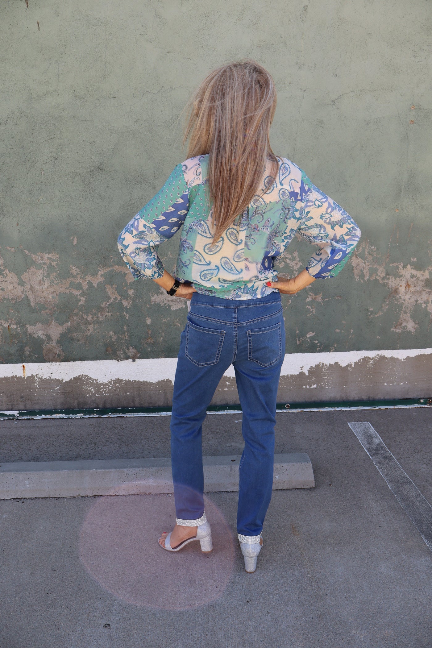 Model is wearing a dark wash blue jean with sparkle details ont the pockets and hemming of ankle. Jeans are straight leg and worn with a paisley blouse and high heels.