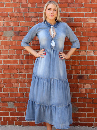 Model is wearing a denim 3/4th sleeve maxi dress with tiered detail, a split neck with a tie at neck, and ruffling at neckline. Worn with converse.