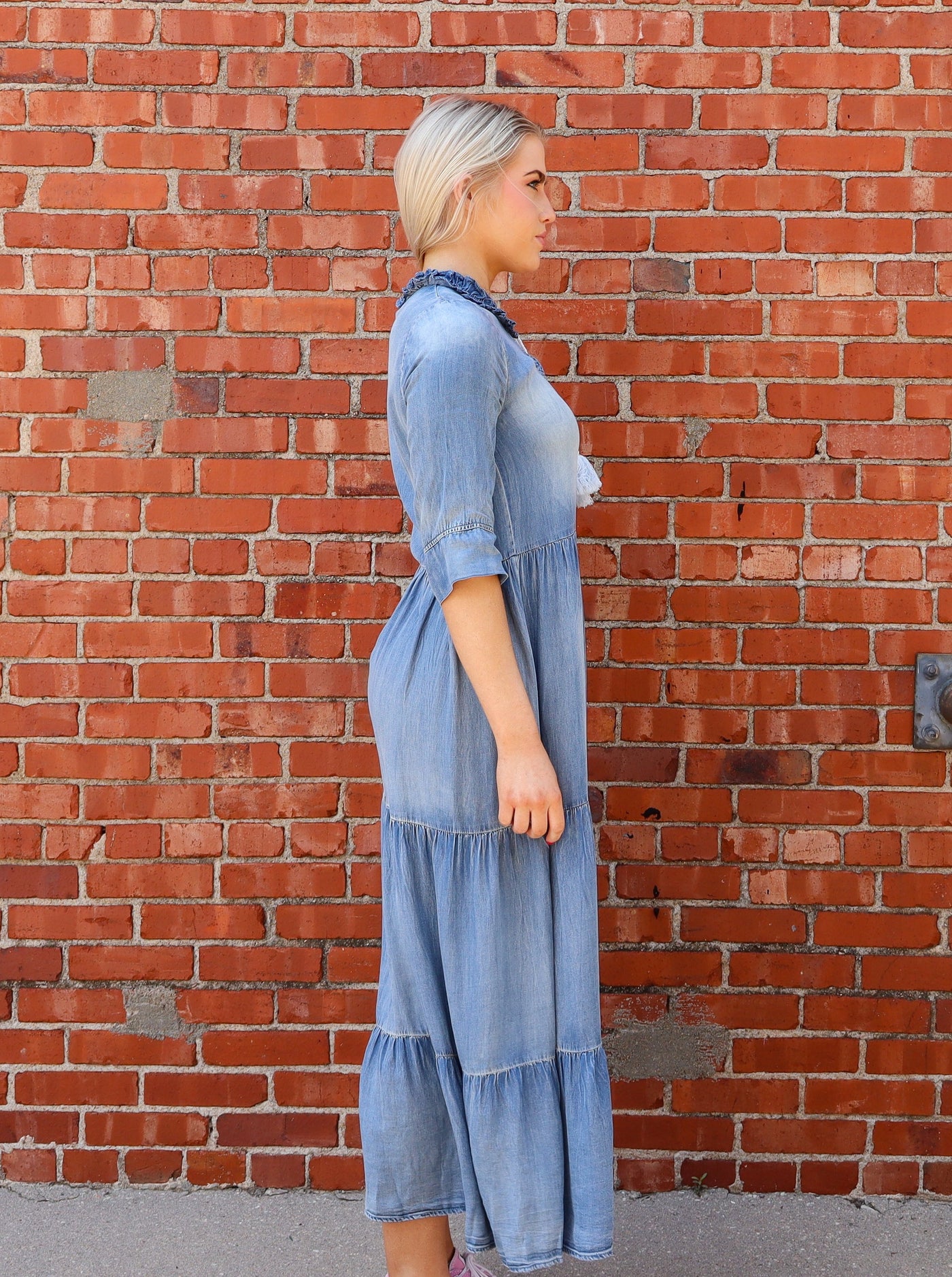 Model is wearing a denim 3/4th sleeve maxi dress with tiered detail, a split neck with a tie at neck, and ruffling at neckline. Worn with converse.