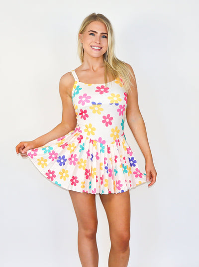 Model is wearing a multi colored flower printed white tennis dress with ruffles at the skirt and a fitted bodice. Worn with white tennis shoes. 