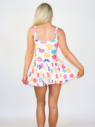 Model is wearing a multi colored flower printed white tennis dress with ruffles at the skirt and a fitted bodice. Worn with white tennis shoes.