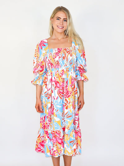 Model is wearing a tropical printed long sleeve maxi dress with a square neckline, ruffles at the sleeves, and tiering with a fitted bodice. 