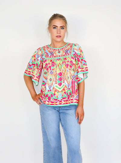 Model is wearing a kaleidoscope theme printed flutter sleeve muti color blouse with a loose fit at the bodice and a round neckline. Worn with blue jeans. 