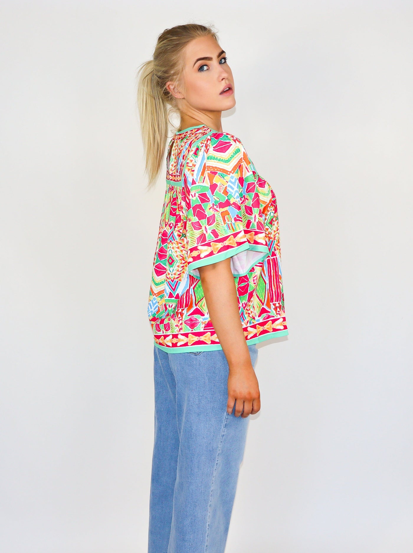 Model is wearing a kaleidoscope theme printed flutter sleeve muti color blouse with a loose fit at the bodice and a round neckline. Worn with blue jeans. 