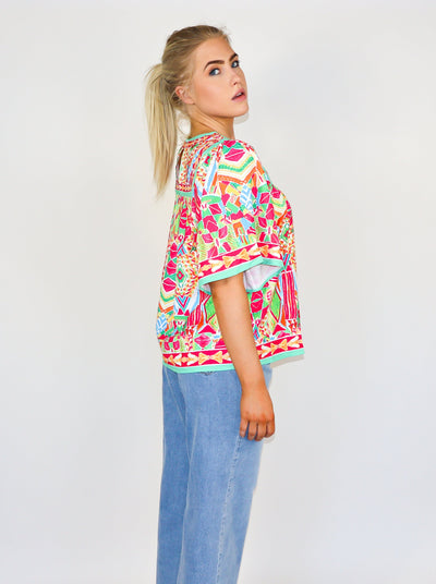 Model is wearing a kaleidoscope theme printed flutter sleeve muti color blouse with a loose fit at the bodice and a round neckline. Worn with blue jeans. 