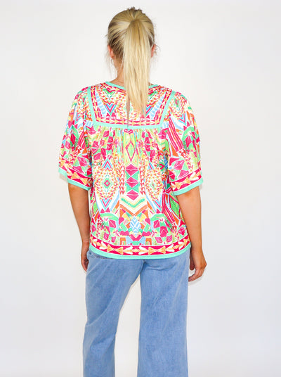 Model is wearing a kaleidoscope theme printed flutter sleeve muti color blouse with a loose fit at the bodice and a round neckline. Worn with blue jeans. 