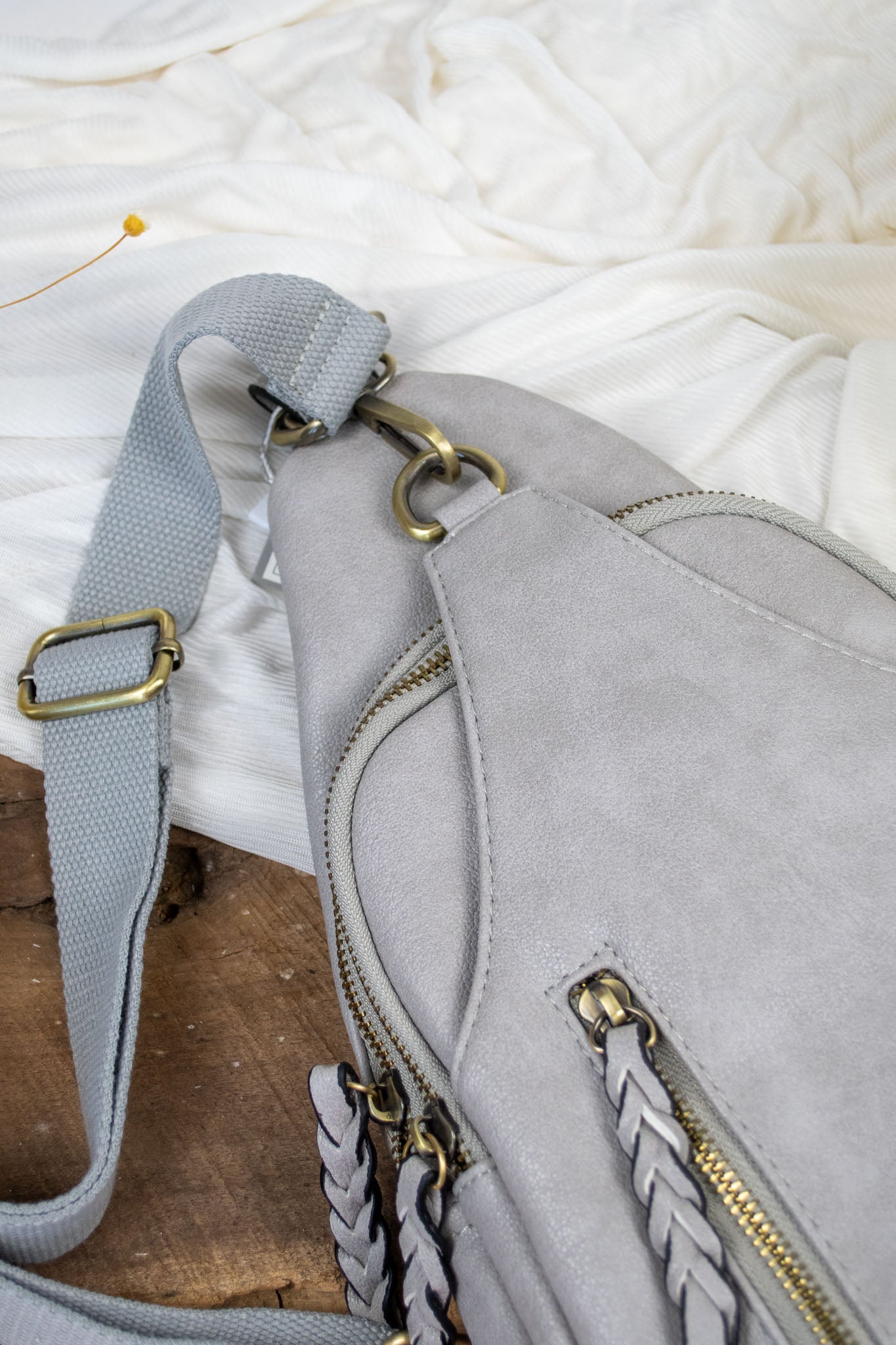A gray vegan leather sling body bag with braided zippers and three compartments.