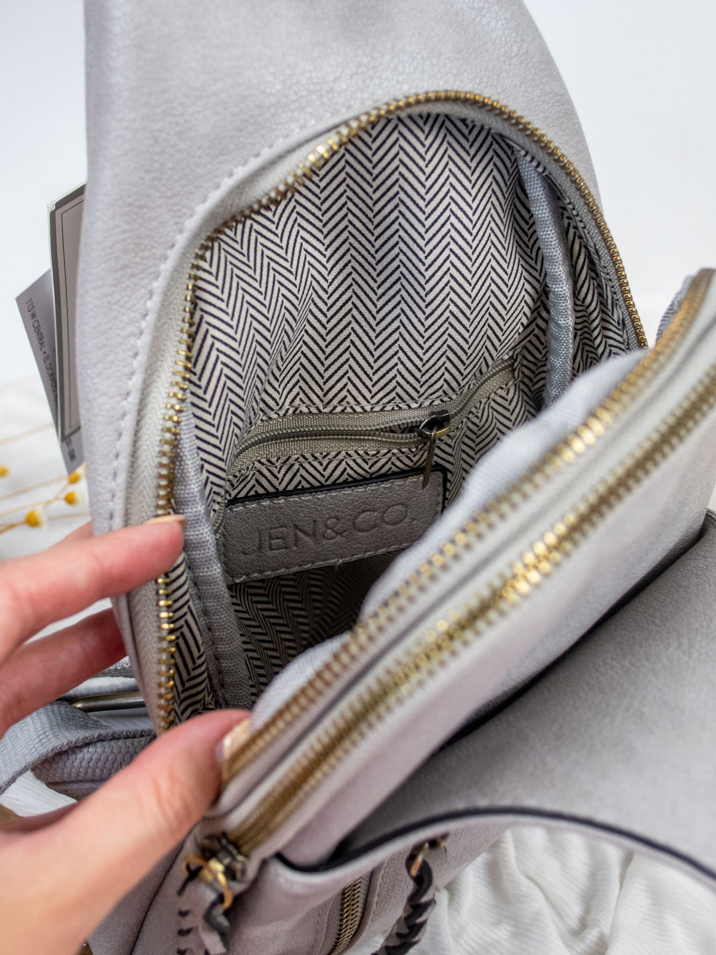 A gray vegan leather sling body bag with braided zippers and three compartments.