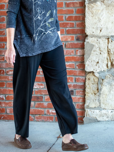 A model wearing a pair of black lantern pants. The model has it paired with a gray floral graphic, asymmetric top and brown loafers. 