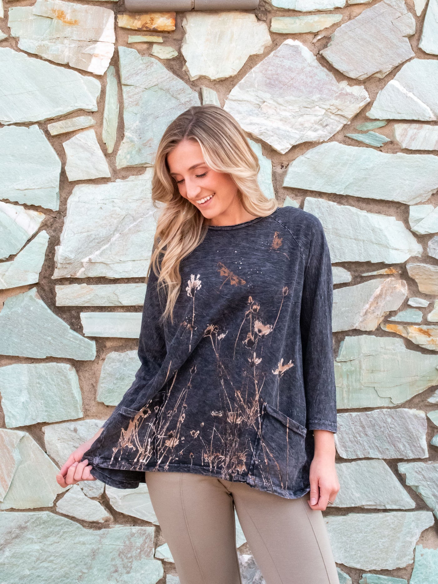 A model wearing a tunic style long sleeve top with a wildflower graphic on the front. The model has it paired with a tan pair of pants.