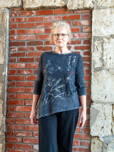 A model wearing a gray asymmetrical top with a floral and dragonfly graphic and 3/4 sleeves. The model has it paired with a pair of black pants.