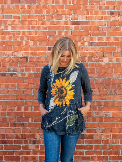 A model wearing a gray, mineral washed, 3/4 sleeve length top with a sunflower graphic. The model has it paired with a pair of medium washed skinny jeans.