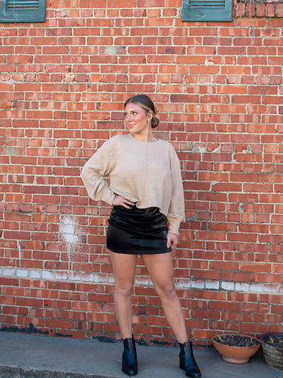 A model wearing a tan sweater with a seam detail. The model has it paired with a black faux leather mini skirt and black booties.