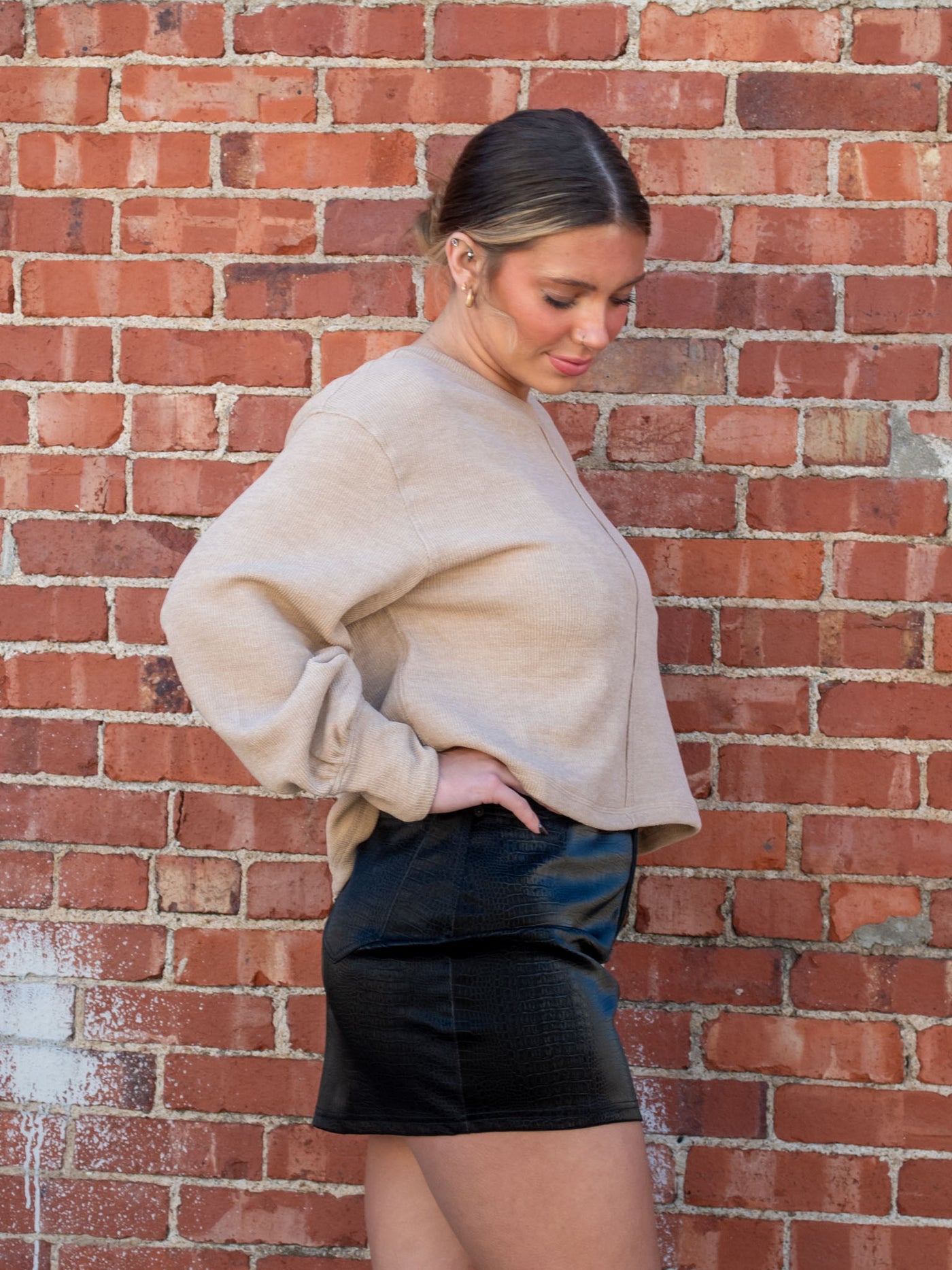 A model wearing a tan sweater with a seam detail. The model has it paired with a black faux leather mini skirt.