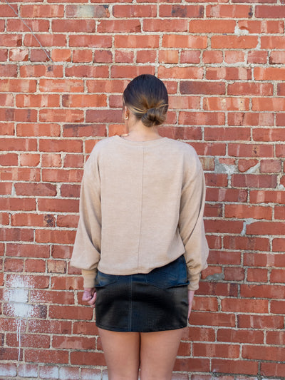 A model wearing a tan sweater with a seam detail. The model has it paired with a black faux leather mini skirt.