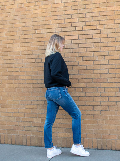 A model wearing a medium wash ankle skinny jean. The model has it paired with a black sweatshirt and white sneakers.